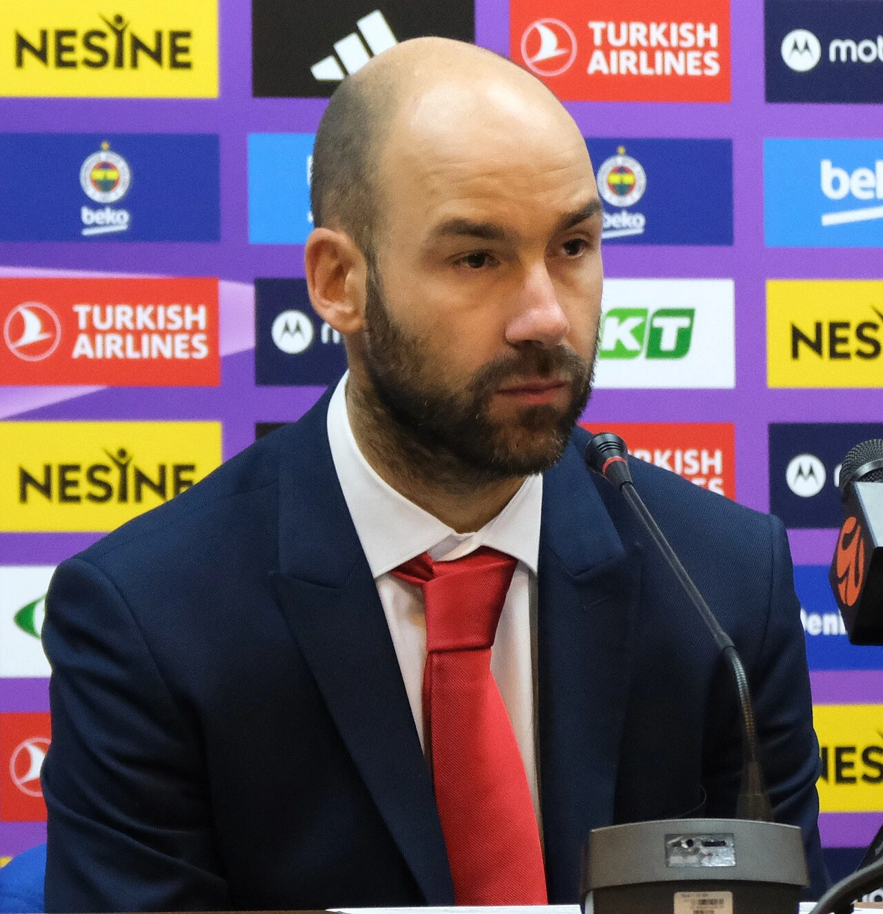 Vassilis Spanoulis speaking at a press conference in a suit and red tie, with sponsor logos in the background.